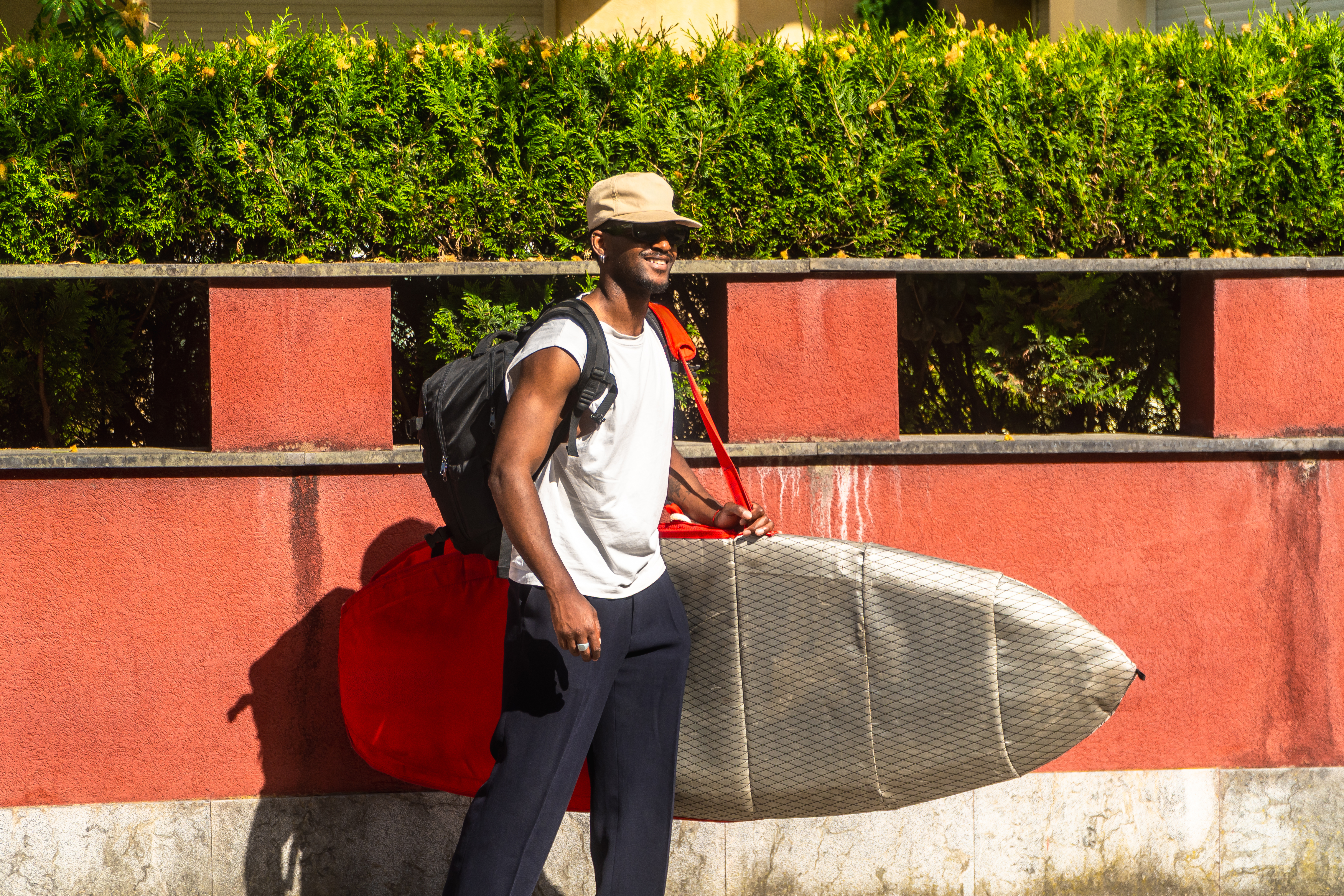 Smiling young adult man carrying a surfboard bag and backpack while walking on a sunny urban street, ready for a surfing adventure